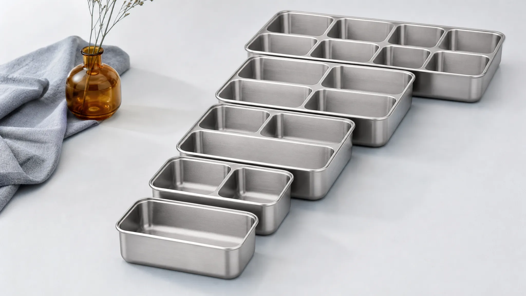 Five brushed stainless steel bento trays of increasing size and compartment count arranged like an ascending staircase