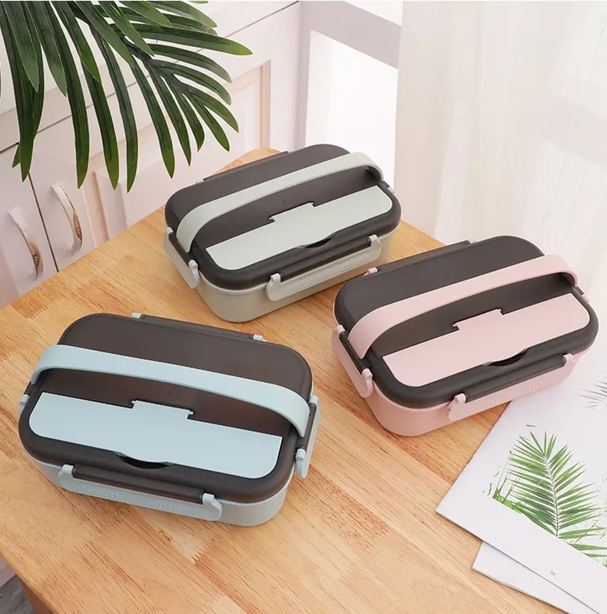 Three children's bento lunch boxes in pale blue, grey and pink with dark lids and silicone band closures, arranged on a light tabletop