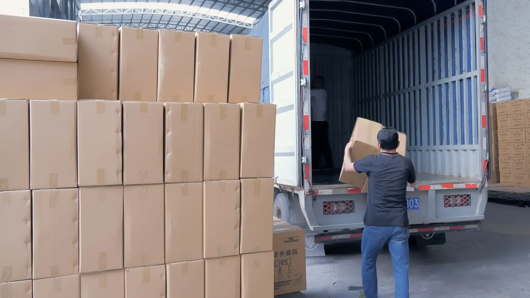 Workers loading sealed kraft cartons into a container truck at a factory loading bay