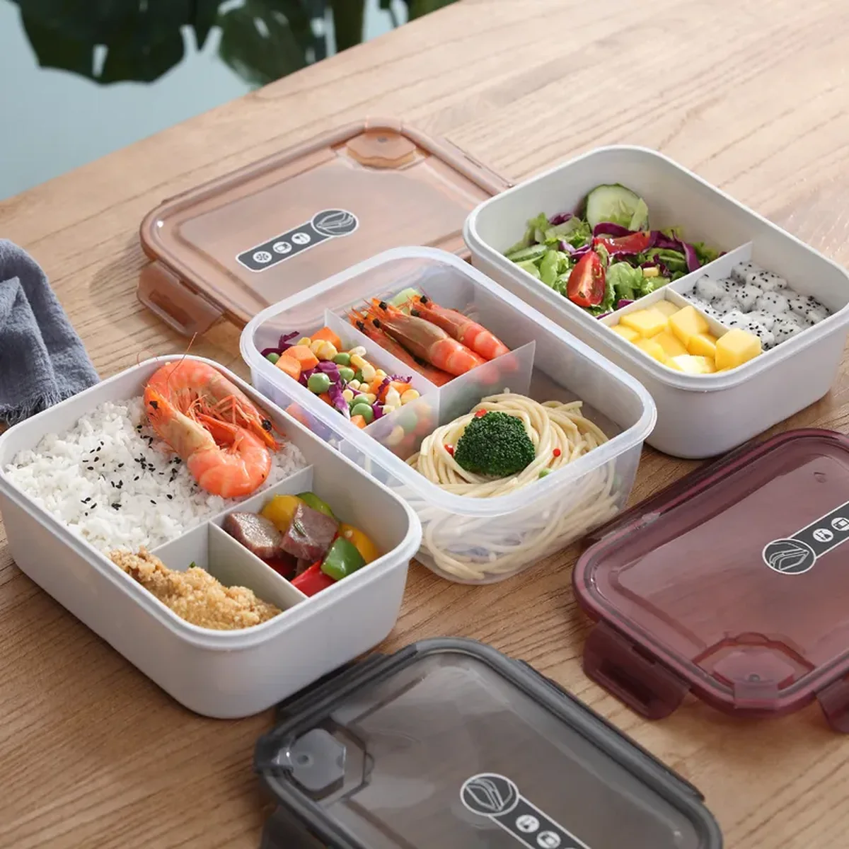 Four clear plastic lunch boxes with three compartments and clipped lids, filled with rice, salad and noodles, on a wooden table