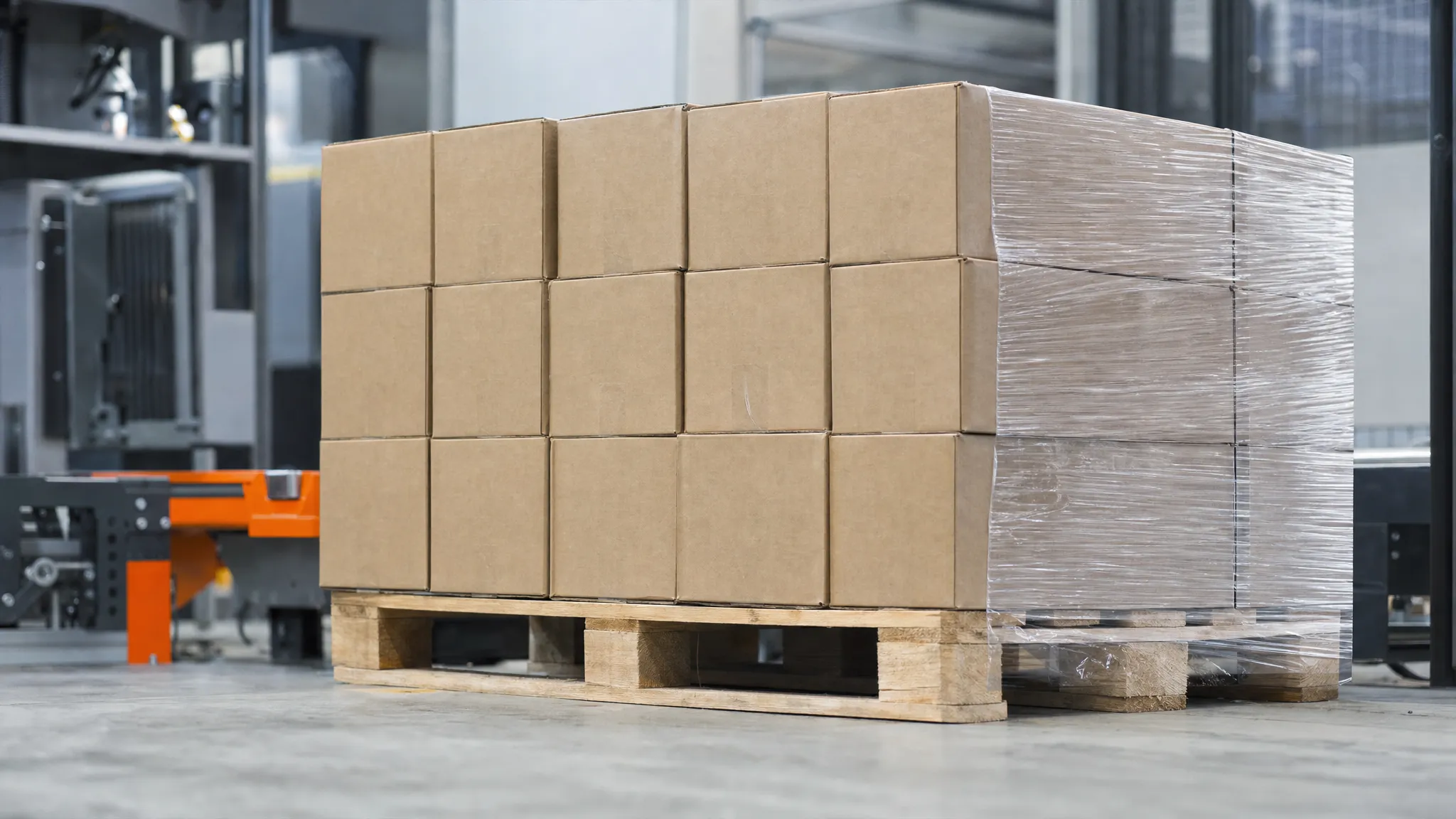 Stacked plain brown export cartons on a wooden pallet in a factory packing area, one corner of the stack shrink-wrapped